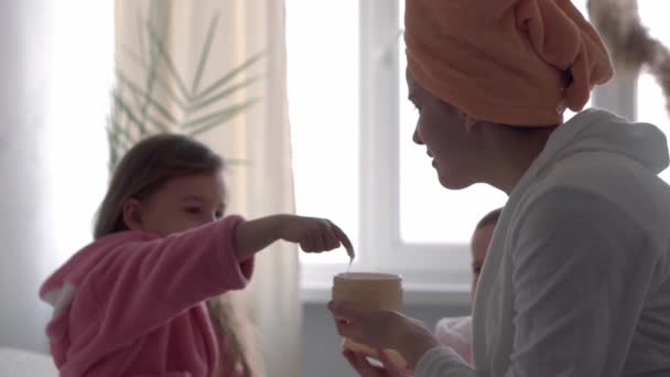 Auténtica linda mujer joven aplicando mascarilla de bebé niña. mamá con 2 hijos hijas de diferentes edades en batas de baño después del baño en la ventana oposite cama blanca. Spa, maternidad, infancia, concepto familiar — Vídeos de Stock