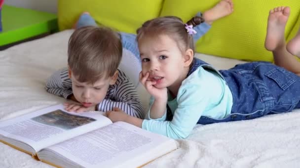 Amistad, familia, educación - Dos preshool sonrientes, niños pequeños leer gran libro interesante de cuentos de hadas en la cama amarilla. Hermanos pequeños gemelos lector divertirse, niños felices en cuarentena en casa — Vídeos de Stock