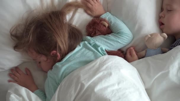 Auténtico retrato lindo caucásico pequeños hermanos preescolares bebé y niña en sueño azul con oso de peluche en la cama blanca. niño descansando a la hora del almuerzo. cuidado, medicina y salud, Infancia, concepto de vida — Vídeo de stock