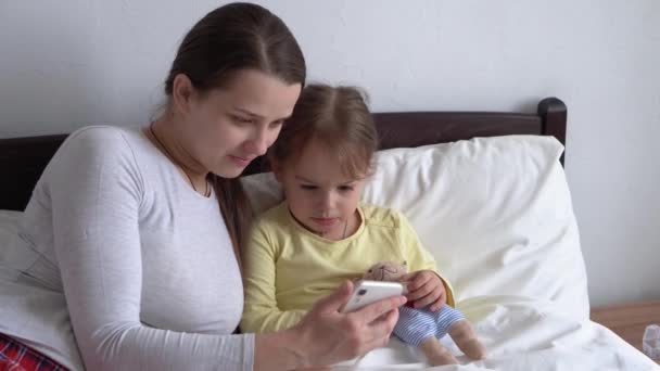 Kaukaska rodzina małe dzieci dziewczynka i młoda kobieta mama rozmawia w wideokonferencji przez smartfona razem w domu z uśmiechem szczęścia na białym łóżku. technologia, styl życia, koncepcja korzystania z telefonu komórkowego — Wideo stockowe