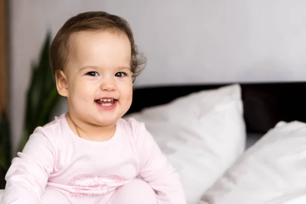 Auténtico retrato caucásico bebé regordete niña o niño en rosa somnoliento al despertar mirando a la cámara sonriendo lindo y muecas en la cama blanca. Cuidado de niños, Infancia, paternidad, concepto de vida —  Fotos de Stock