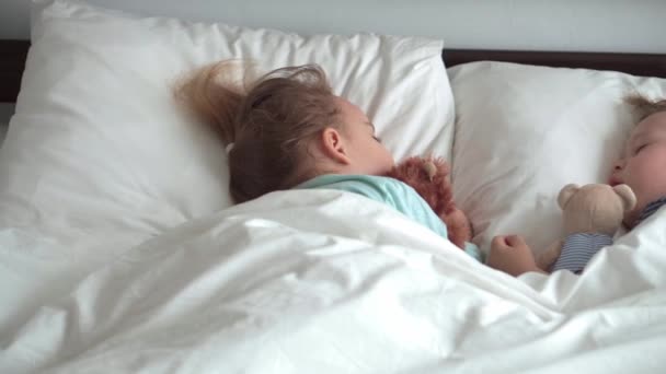 Auténtico retrato lindo caucásico pequeños hermanos preescolares bebé y niña en sueño azul con oso de peluche en la cama blanca. niño descansando a la hora del almuerzo. cuidado, medicina y salud, Infancia, concepto de vida — Vídeo de stock