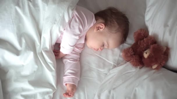 Authentisches Porträt niedlichen kaukasischen kleinen Säugling pummelig Baby Mädchen oder Junge im rosa Schlaf mit Teddybär auf weißem Bett. Mittags ruhen die Kinder. Pflege, Schlafkind, Kindheit, Elternschaft, Lebenskonzept — Stockvideo