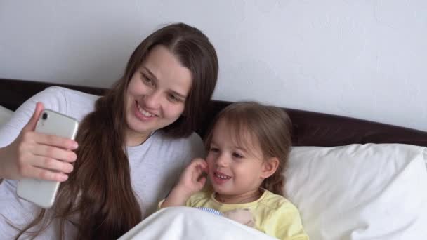 Kaukaska rodzina małe dzieci dziewczynka i młoda kobieta mama rozmawia w wideokonferencji przez smartfona razem w domu z uśmiechem szczęścia na białym łóżku. technologia, styl życia, koncepcja korzystania z telefonu komórkowego — Wideo stockowe