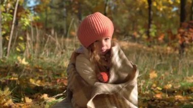 Otantik, küçük, beyaz, beyaz, 3-4 yaş arası, turuncu bereli bir kız çocuğu. Sonbahar parkında ya da ormanda yere düşmüş sarı yapraklara sarılmış bir battaniyeye oturuyor. Doğa, Mevsim, Çocukluk kavramı.