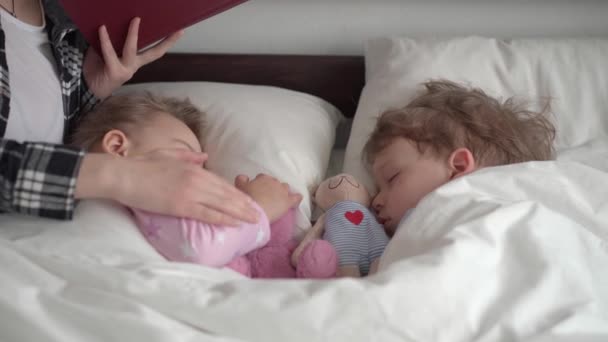 Auténtica joven linda mujer o niñera lee cuento de hadas de libro a los niños antes de acostarse. Mamá se encarga de los niños dormidos. Dos bebés durmiendo en una cama blanca. Día de la madre, maternidad, infancia — Vídeo de stock