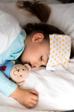 Otantik portre, beyaz yatakta alnına baskı uygulayan mavi elbiseli, beyaz, beyaz, şirin, beyaz bir anaokulu bebeği. Çocuk öğle yemeği vakti dinleniyor. bakım, ilaç ve sağlık, çocukluk, yaşam konsepti