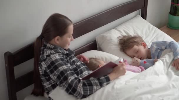 Authentic cute young woman or babysitter reads fairy tale from book to children before bedtime. Mom takes care of sleeping children. Two baby sleeping in white bed. Mothers day, motherhood, childhood — Stock Video