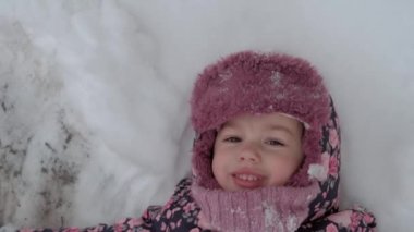 Tatil, oyun, çocukluk konsepti - yavaş çekim, otantik, mutlu, anaokulu çocuğu bebek gülümsemesi karların üzerinde yatar ve ayaklarla alkış tutarak melek yapar. Soğuk mevsimde kar yağışı Kışın açık havada