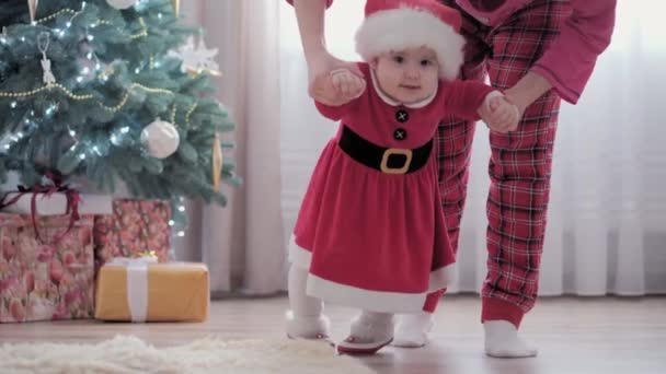Подлинный милый пухлый младенец девочка в шляпе Санта-Клауса и красное платье учится ходить делает первые шаги с молодой мамой празднуя Новый год рядом с елкой. Детство, материнство, уход, зимняя концепция. — стоковое видео