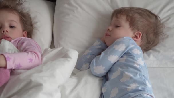 Authentieke Kaukasische 2 kleine broertjes en zusjes peuters peuter meisje jongen in roze blauw slaperig bij het wakker worden kijkend naar camera lachend schattig en grimmig in wit bed. Kinderopvang, Kindertijd, Ouderschap, Levensstijl — Stockvideo