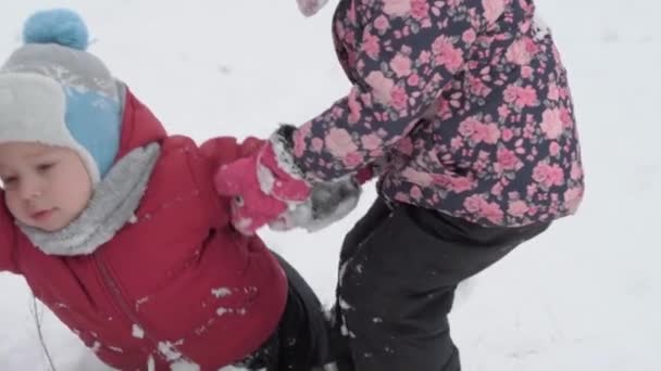 休暇、ゲーム、家族の概念- slo-mo本物の2つの幸せな就学前の幼児の兄弟姉妹は丘を滑り落ち、互いに登るのを助けます。冬の公園の寒い季節の天気の降雪屋外 — ストック動画