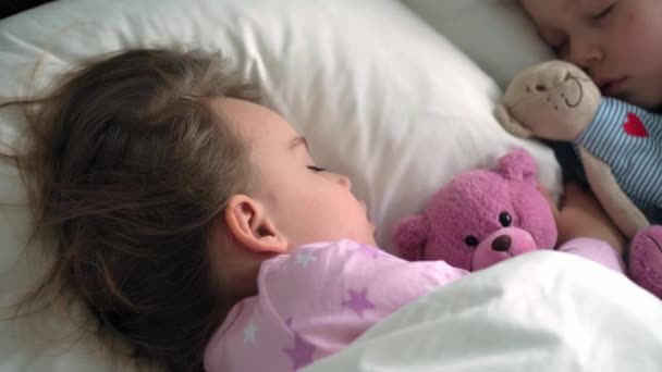 Auténtico retrato lindo caucásico pequeños hermanos preescolares bebé y niña en pijama rosa azul dormir con oso de peluche en la cama blanca. descanso infantil, cuidado, medicina y salud, Infancia, concepto de vida — Vídeo de stock