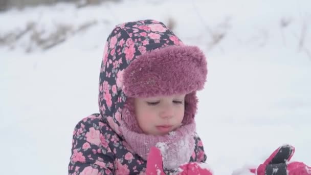 Roeping, spel, kindertijd concept - slo-mo authentieke vrolijke kleuter peuter baby meisje met hoed en wanten likken sneeuwvlokken van lippen en eet sneeuw. sneeuwval in het koude seizoen weer in de winter buiten — Stockvideo