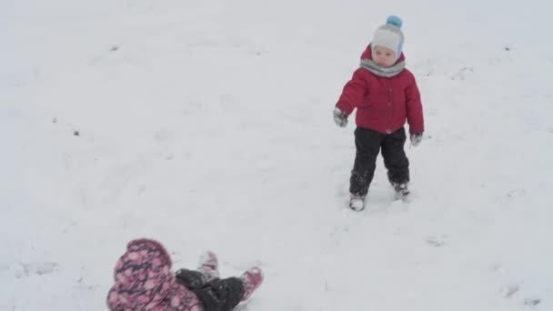 Wakacje, gra, koncepcja rodziny - slo-mo autentyczne Dwa szczęśliwe przedszkolaki rodzeństwo dzieci zjeżdżają ze wzgórza i pomagają sobie nawzajem wspiąć. opady śniegu w zimnym sezonie pogoda w parku zimowym na zewnątrz — Wideo stockowe