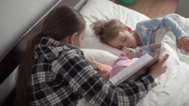 Auténtica joven linda mujer o niñera lee cuento de hadas de libro a los niños antes de acostarse. Mamá se encarga de los niños dormidos. Dos bebés durmiendo en una cama blanca. Día de la madre, maternidad, infancia — Vídeos de Stock