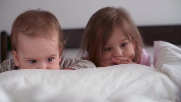 Autentikus 3 kis óvodás kisgyerek kisgyermekek testvérek nézni rajzfilm okostelefon eszköz együtt baba fehér ágy otthon beltérben. Család, szabadidő, gyermekkor, barátság koncepció. — Stock videók