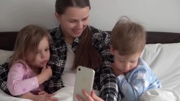 Authentique jeune femme mère de nombreux enfants avec bébé garçon et fille parlant en vidéoconférence par smartphone ensemble à la maison avec le sourire sur lit blanc. technologie, mode de vie, concept d'utilisation du téléphone mobile — Video