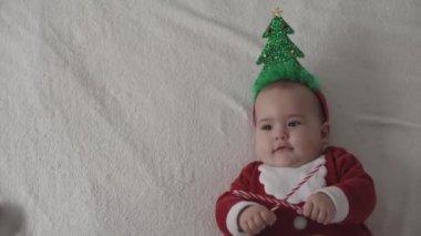 Mutlu Noeller, mutlu yıllar, bebekler, çocukluk, tatil konsepti - yakın plan gülümseyen 7 aylık yeni doğan bebek Noel ağacı çemberinde, kırmızı tulum beyaz yumuşak yatakta yatıyor tatlı şekerlemeleri yala