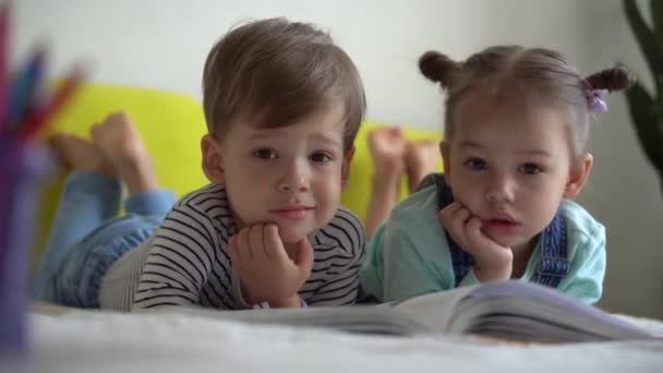 Amistad, familia, educación - Dos preshool sonrientes, niños pequeños leer gran libro interesante de cuentos de hadas en la cama amarilla. Hermanos pequeños gemelos lector divertirse, niños felices en cuarentena en casa — Vídeos de Stock