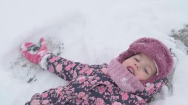 Tatil, oyun, çocukluk konsepti - yavaş çekim, otantik, mutlu, anaokulu çocuğu bebek gülümsemesi karların üzerinde yatar ve ayaklarla alkış tutarak melek yapar. Soğuk mevsimde kar yağışı Kışın açık havada