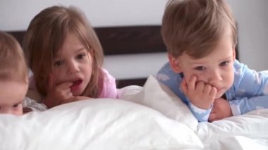 Otantik 3 küçük anaokulu çocuğu reşit olmayan çocuklar, kardeşler akıllı telefon cihazıyla çizgi film izliyorlar evde beyaz yatakta bebek gibi. Aile, eğlence, çocukluk, arkadaşlık konsepti..