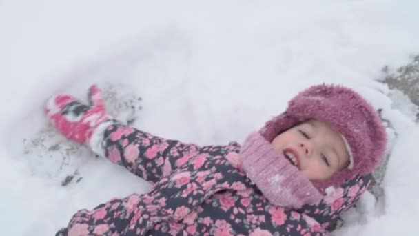 Vacanza, gioco, concetto di infanzia - Slo-mo autentico felice bambino in età prescolare sorriso bambina si trova nella neve sul retro e applaudire con i piedi fanno angelo. nevicate nella stagione fredda tempo in inverno all'aperto — Video Stock