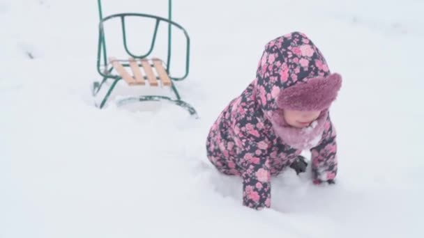 Slo-mo auténtica niña preescolar feliz en sombrero y manoplas se arrastran a cuatro patas en la nieve y cae sobre la espalda. nevadas en temporada fría en invierno al aire libre. vocación, juego, concepto de infancia — Vídeos de Stock