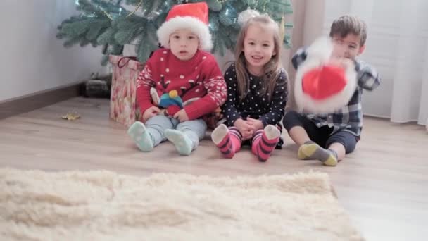 Tre autentiska glada glada Joy barn sitta vid dekorerad julgran leka ha kul tillsammans. Förskolebarn firar nyår festlig atmosfär hemma. Vinter, semester, Familjebegreppet — Stockvideo