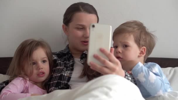 真正的年轻女性母亲带着许多孩子，带着男婴和女婴在视频会议上用智能手机在家里一起聊天，笑着躺在白色的床上。技术、生活方式、移动电话使用概念 — 图库视频影像