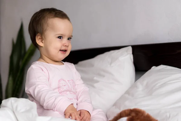 Auténtico retrato caucásico bebé regordete niña o niño en rosa somnoliento al despertar mirando a la cámara sonriendo lindo y muecas en la cama blanca. Cuidado de niños, Infancia, paternidad, concepto de vida — Foto de Stock