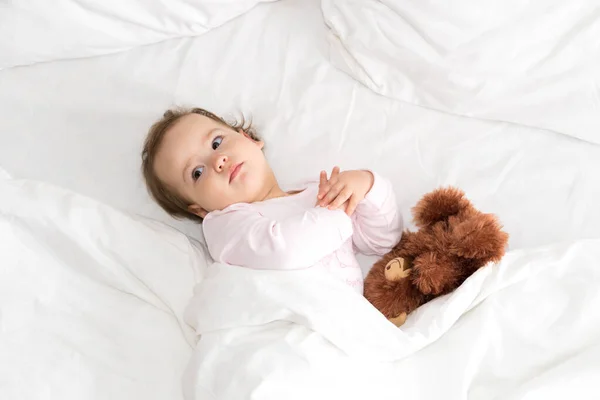 Retrato autêntico bonito caucasiano pequeno bebê gordinho menina ou menino em rosa sonolento ao acordar com ursinho olhando para a câmera na cama branca. Cuidados infantis, Infância, Paternidade, conceito de estilo de vida — Fotografia de Stock