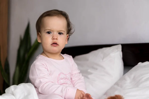 Retrato autêntico bonito caucasiano pequeno bebê gordinho menina ou menino em rosa sonolento ao acordar olhando para a câmera na cama branca. Cuidados infantis, Infância, Paternidade, conceito de estilo de vida — Fotografia de Stock