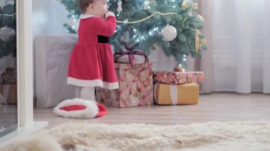 Noel Baba şapkası takan, kırmızı elbiseli, mutlu, mutlu, tombul bir kız çocuğu. Yeni yıl atmosferini kutlarken evdeki Noel ağacının yanında iyi eğlenceler. Çocukluk, bayram, kış konsepti
