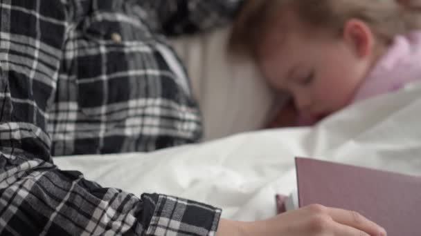 Autentyczne słodkie młoda mama zasnęła czytać bajki z książki do dziecka przed snem. Kobieta zajmuje się śpiącymi dziećmi. Dwójka dzieci śpi w białym łóżku. Dzień Matki, macierzyństwo, koncepcja dzieciństwa — Wideo stockowe