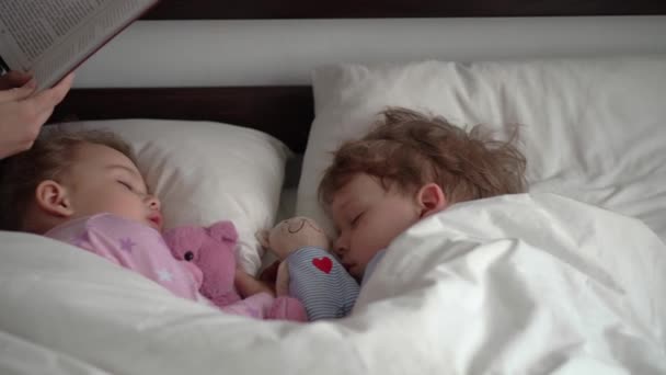 Authentique jeune femme mignonne ou baby-sitter lit conte de fées du livre aux enfants avant le coucher. Maman s'occupe des enfants endormis. Deux bébés dormant dans un lit blanc. Fête des mères, maternité, enfance — Video