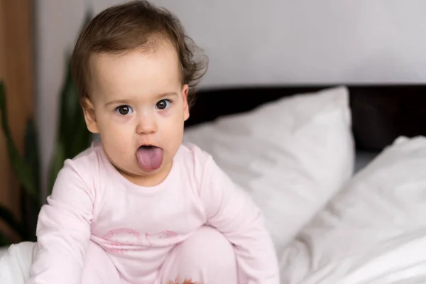 Retrato autêntico bonito caucasiano pequeno bebê gordinho menina ou menino em rosa sonolento ao acordar olhando para a câmera mostrando a língua na cama branca. Cuidados infantis, Infância, Paternidade, conceito de estilo de vida — Fotografia de Stock