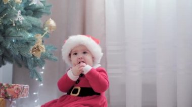 Noel Baba şapkası takan, kırmızı elbiseli, mutlu, mutlu, tombul bir kız çocuğu. Yeni yıl atmosferini kutlarken evdeki Noel ağacının yanında iyi eğlenceler. Çocukluk, bayram, kış konsepti