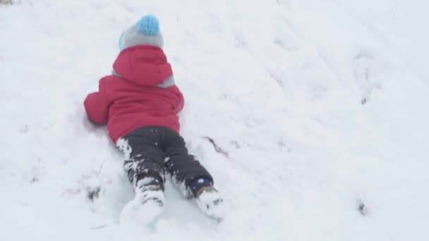 Vacation, game, family concept - slo-mo authentic One happy preschool toddler kid child boy in warm hat sliding on stomach belly from snow from hill. snowfall in cold weather in winter park outdoors — Stock Video