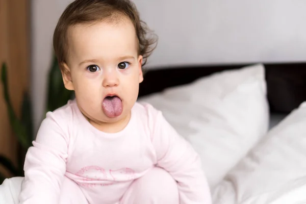 Retrato autêntico bonito caucasiano pequeno bebê gordinho menina ou menino em rosa sonolento ao acordar olhando para a câmera mostrando a língua na cama branca. Cuidados infantis, Infância, Paternidade, conceito de estilo de vida — Fotografia de Stock