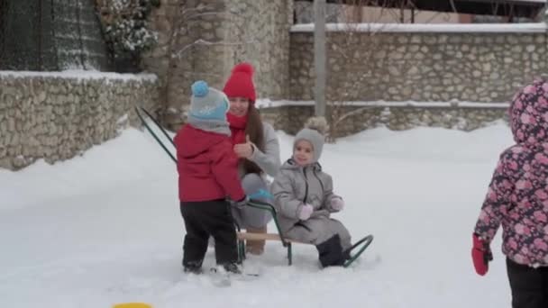 Відпустка, гра, сімейна концепція - шлю-мо автентичні три щасливі діти дошкільного віку брати і сестри з молодою мамою, що катаються на санчатах і граються зі снігом. снігопад в холодну погоду в зимовому дворі на відкритому повітрі — стокове відео