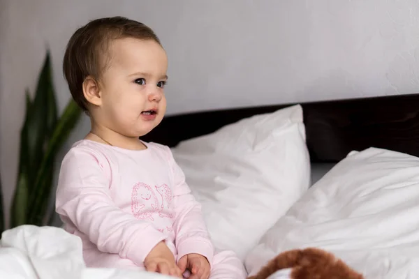 Retrato autêntico bonito caucasiano pequeno bebê gordinho menina ou menino em rosa sonolento ao acordar olhando para a câmera na cama branca. Cuidados infantis, Infância, Paternidade, conceito de estilo de vida — Fotografia de Stock