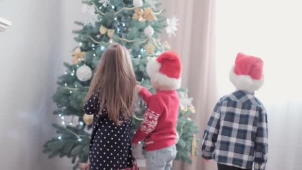 3 niños felices auténticos alegría saludar de la mano regocijarse por el juego del árbol de Navidad divertirse juntos. Los niños preescolares celebran el ambiente festivo de Año Nuevo en casa. Invierno, vacaciones, concepto familiar — Vídeo de stock