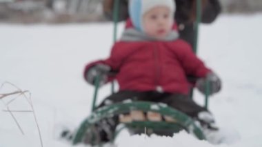 Tatil, oyun, aile kavramı - yavaş çekim, otantik, mutlu, mutlu, gülümseyen genç anne yeni yürümeye başlayan bir anaokulu çocuğuyla kızak sürüyor, ablası genç kardeşiyle kızak kayıyor kışın karlı ve soğuk havada açık havada.