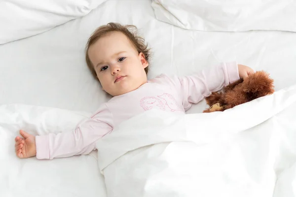 Auténtico retrato lindo bebé caucásico regordete niña o niño en rosa somnoliento al despertar con oso de peluche mirando a la cámara en la cama blanca. Cuidado de niños, Infancia, paternidad, concepto de estilo de vida — Foto de Stock