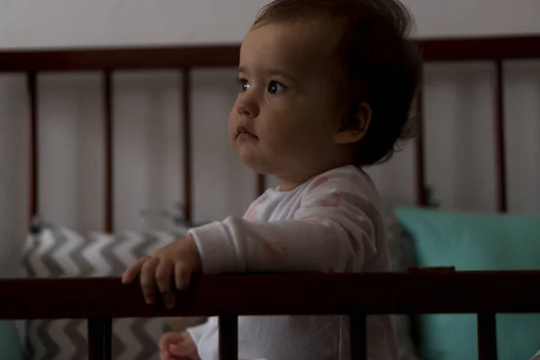 Linda niña regordeta alegre sentada en una cuna gris después de despertar de dormir en pijamas blancos y rosados con oso de peluche y sonriente. Infancia, ocio, comodidad, medicina, concepto de salud — Foto de Stock