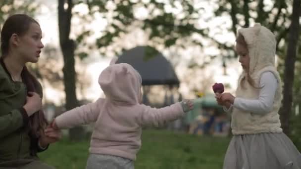 Jovem mãe recolhe flores de dente-de-leão amarelo com dois irmãos bebês irmãs menina no parque da primavera. Feliz mãe bonita crianças Brincar ter diversão cheiro tulipas ao pôr do sol. Infância Lazer Natureza do tempo — Vídeo de Stock