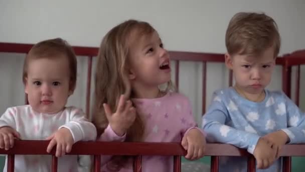 Leuke vrolijke 3 kleine kinderen meisje en jongen broers en zussen kleuterschool kinderen in grijze babybedje na het wakker worden in een pyjama met benen bungelend. Jeugd, vrije tijd, comfort, geneeskunde, gezondheidsconcept — Stockvideo