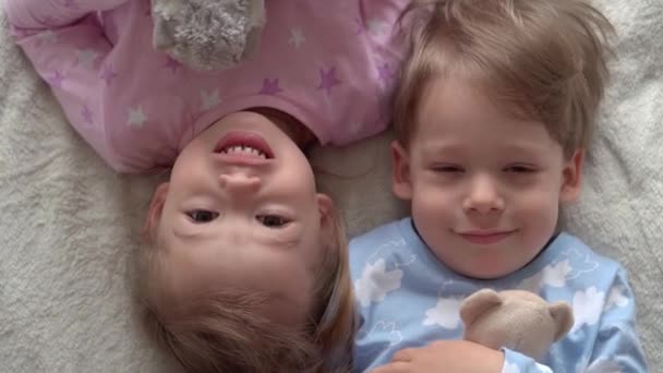 Dos niños sonrientes de preescolar niño y niña tumbados en la espalda miran en la cámara en pijama en la cama. Los bebés gemelos se divierten, niños felices en cuarentena en casa. Amistad, familia, concepto educativo — Vídeos de Stock