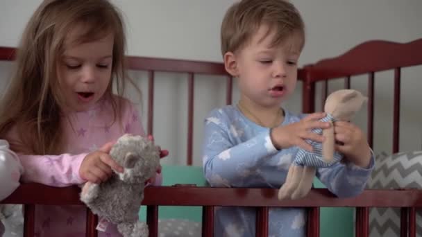 Cute cheerful 3 little kids girl and boy siblings preschool children in grey baby crib after waking up from sleep in pajamas with legs dangling. Childhood, leisure, comfort, medicine, health concept — Stock Video